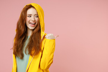 Redhead young woman in yellow waterproof raincoat outerwear hood point thumb finger aside on...