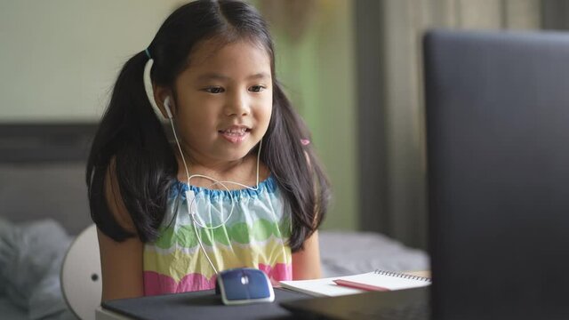 Asian child student or kid girl smile e-learning on computer notebook to wearing headphone for video call communication by enjoy study online or people learn from home to play laptop by back to school
