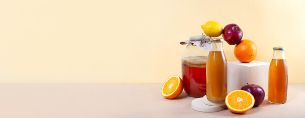 Banner. Kombucha tea in glass jar, two bottles and balancing fruits for additional flavors on podium on pastel background. Orange, apple and lemon balance on modern pedestal. Healthy fermented drink
