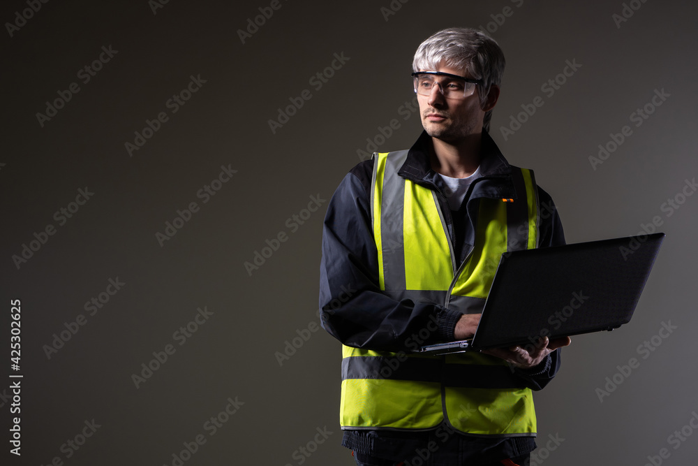Poster man in yellow vest holds a laptop. concept - he's computer scientist in a industrial factory. career