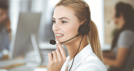 Blonde business woman using headset for communication and consulting people at customer service office. Call center. Group of operators at work