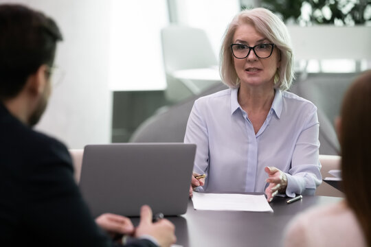 Confident middle-aged Caucasian businesswoman talk with colleagues or clients at office meeting. Successful mature female leader or boss lead head briefing with business partners. Leadership concept.