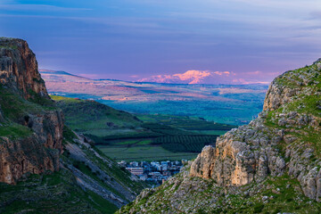 Beautiful view of Galilee from the cliff of Mount Arbel National Park and Nature Reserve, with the snow-capped Mt Hermon lit up by pink sunset light in the distance; Lower Galilee, Israel - obrazy, fototapety, plakaty