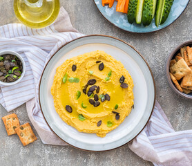 Healthy snack, chickpea hummus with pumpkin or carrots and pumpkin seeds on a ceramic plate on a gray concrete background. Served with savory crackers.