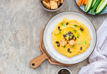 Healthy snack, chickpea hummus with pumpkin or carrots and pumpkin seeds on a ceramic plate on a gray concrete background. Served with savory crackers.