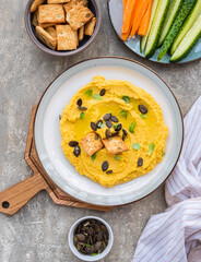 Healthy snack, chickpea hummus with pumpkin or carrots and pumpkin seeds on a ceramic plate on a gray concrete background. Served with savory crackers.