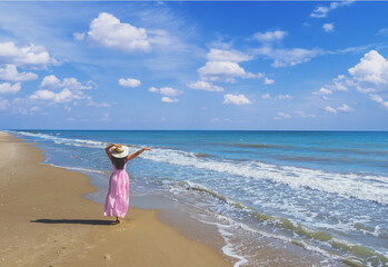 Fototapeta na wymiar Seascape with beautiful sky. Woman on the beach, summertime. Young happy woman in a long pink dress and straw hat walking barefoot on the seaside. View from above