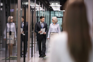 Diverse businesspeople walk down modern office hallway talking or chatting. Casual life or day...