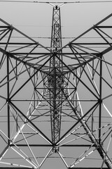 Black and white photo standing exactly under an electricity pole looking from bottom to top with symmetrical shapes of the steel structure.