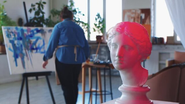 A woman paint artist drawing on a canvas with blue paint - greek head sculpture on the foreground in red neon lighting