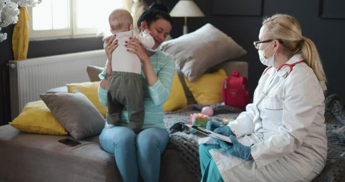 Visiting Patronage Nurse Visiting A Young Mother And A Baby At Home. Portrait Of Young Mom In Mask Holding Cute Baby And Listening To Doctor During Medical Checkup At Home. Informing Young Mothers