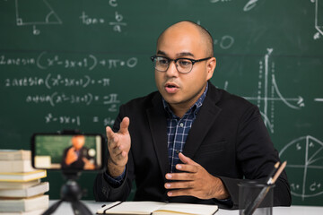 Indian young teacher man sitting teaching online video conference live stream by smartphone. Asian teacher teaching mathematics class webinar online for students learning.