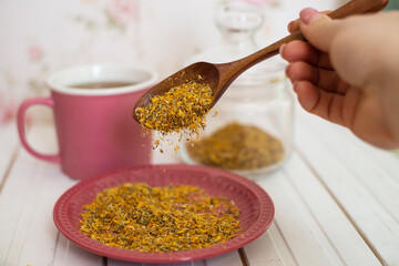 Arnica dried flowers in the wooden spoon falling down onto pink plate, the cup of tea and glass jar...