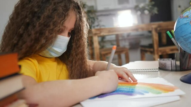 child kid in a mask fun draws a rainbow at home. stay at home coronavirus concept. girl kid in mask draws a rainbow at kindergarten. Kid rainbow drawing coronavirus. stay home