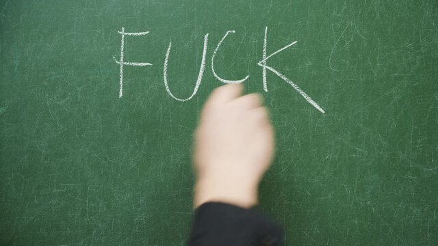 Male hand writing words fuck school with chalk on blackboard or green chalkboard in classroom