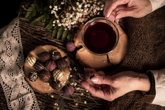 Teezeit in rustikal Still. Schleier Kraut liegt als Deko auf dem Holztisch. Glas Tee steht auf dem Holzhintergrund, bunte Pralinen liegen liegen auf dem St&uuml;ck Holz. H&auml;nde einer Frau, 