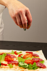 female hand sprinkles fresh chopped greens on unwrapped shawarma with chopped vegetables and salami sausage on black wooden table