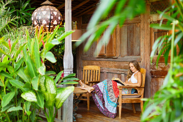 Attractive girl sit on open-air veranda of wood house with tropical garden view, read romance in paper book. Young woman relaxing in luxury villa on family summer vacation. Tropical island lifestyle. - obrazy, fototapety, plakaty