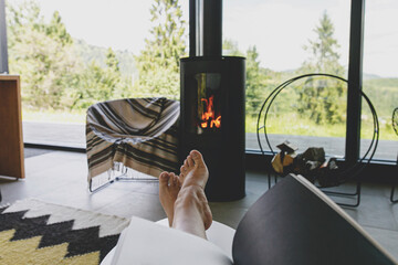 Beautiful female feet and book on background of warm fireplace and big window in modern cozy chalet