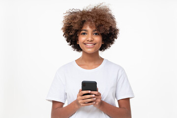 Fototapeta na wymiar African american smiling teenage girl with afro hairstyle holding phone with both hands, chatting with friend, using social media app, isolated on gray background