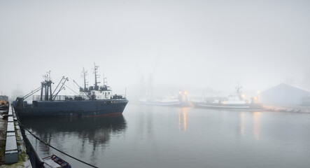 Tugboats and fishing boats (trawlers) moored to a pier in a harbor. Thick white fog. Latvia, Baltic...