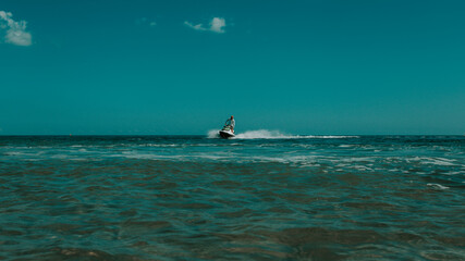 Naklejka premium A man on a jet ski ride, the sea and the ocean in clothes, hot weather with the rays of the bright sun and white clouds against the background of the blue sky and blue transparent water with splashes