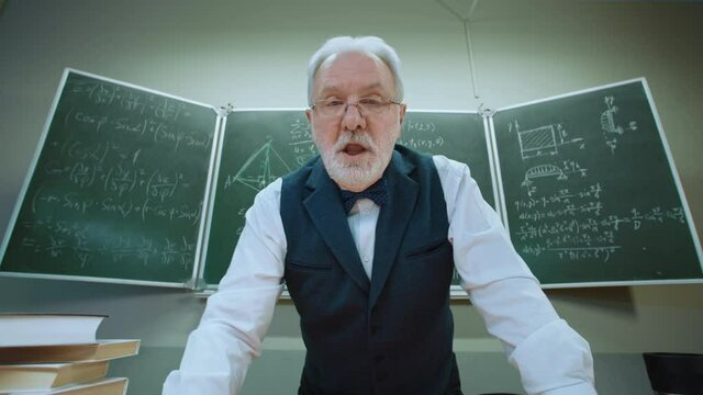 Emotional crazy teacher trying explain mathematical formulas written in chalk on blackboard in classroom. POV view funny senior professor emotionally speaking to student who does not understand math