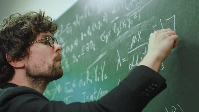Intelligent stylish teacher wearing eyeglasses writes math formulas with chalk on green chalkboard in university or school classroom. Education and studying concept - Powered by Adobe