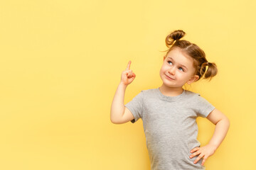 Portrait of child girl with finger pointed up. Little kid in pose has idea isolated on yellow...