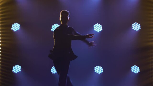 A silhouette of man dancer who is dancing rumba elements. Ballroom dancer is practicing Latin American dances on the parquet in a dark studio with smoke and lights. Close up.
