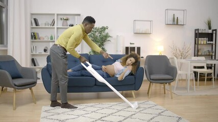Caring husband vacuuming the floor, pregnant wife relaxing on sofa, household - Powered by Adobe