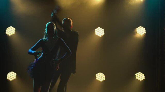 Rumba drive dance performed by a young pair of ballroom dancers. Man in suit and woman in short dress are dancing merrily in a dark studio against a background of bright lights. Silhouettes. Close up.