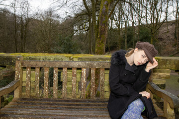 A attractive young woman in a public park sat on an old bench on a cold day in the winter time looking upset and scared