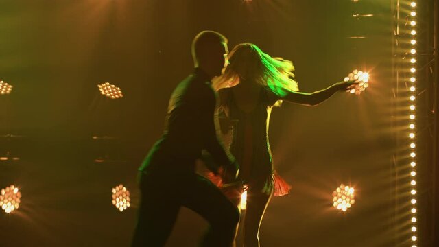 Passionate blonde is dancing rumba with her partner against backdrop of colorful lights. Silhouettes of couple dancers performing Latin American ballroom dance elements in a dark studio. Close up.