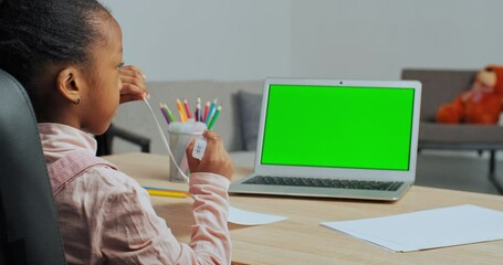 Back view of afro american schoolgirl little ethnic girl sitting at home at table draws picture with colored markers looks into green screen of laptop, online lesson, remote learning in quarantine - Powered by Adobe