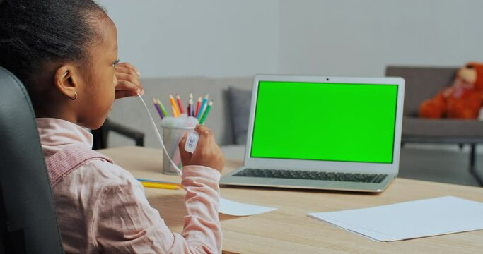 Back view of afro american schoolgirl little ethnic girl sitting at home at table draws picture with colored markers looks into green screen of laptop, online lesson, remote learning in quarantine