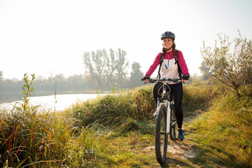 Happy young Caucasian woman riding mountain bike along the footpath through tall grass on the river bank in early morning sunshine