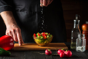 Chef sprinkles salt salad of fresh vegetables on wooden table. Preparing healthy food on the kitchen
