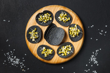 Black nori seaweed chips with mung bean sprouts on a round wooden tray. Top view on a black background, with grains of sea salt.