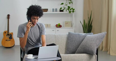Serious afro american business man student guy talking on mobile phone use laptop for remote work studying from home office. Freelancer consults by smartphone communicates online in apartment interior - Powered by Adobe