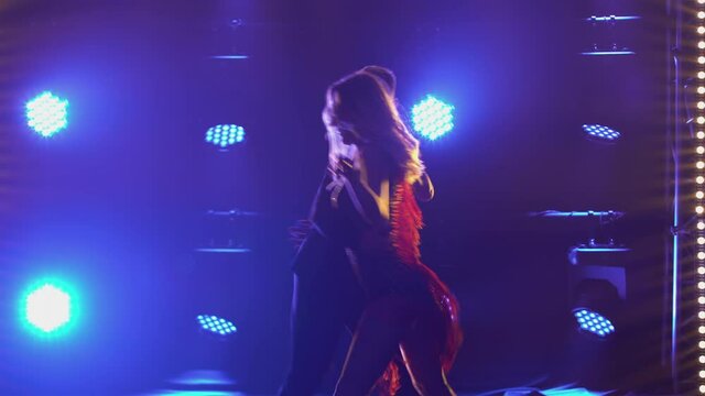 Incendiary rumba performed by ballroom dancing partners. A man in a suit and a woman in a chic short dress dance passionately in a dark studio with bright blue lights. Silhouettes. Close up.