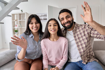 Happy indian couple and teen child daughter waving hands looking at camera sitting on couch at...