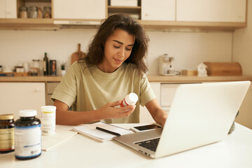 Beautiful young female dietician working from home, using portable computer, typing article about vitamins, holding bottle of food supplements, reading label on it. Health, wellness and dieting