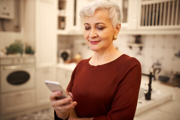 Fototapeta na wymiar Joyful retired female with gray hair holding smart phone reading motivational post or text message while messaging with friend via online application. Mature lady enjoying using electronic gadget