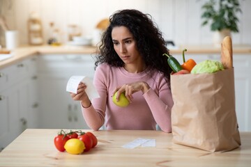 Concerned Young Woman Checking Bills In Kitchen After Grocery Shopping
