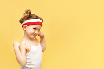 Excited fitness child posing in sportswear,red headband over yellow studio background. Children sport,advertising,shopping concept.Gym workout.Childhood activity.Sport.Health and energy