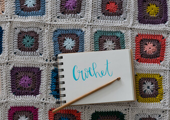 Blue word "Crochet" written in an empty spiral notebook over a granny square blanket. Horizontal image.