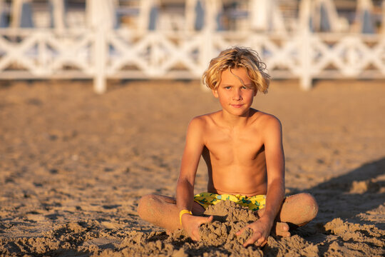 Cute blond teen boy in yellow swim shorts sitting at the sand beach and playing with the sand against white hotel fence. Copy space.