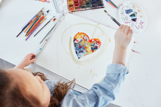 Cute child is drawing colorful heart on white paper. Art school concept. Top view of a girl kid sitting at the table and painting with watercolor paints. High quality photo