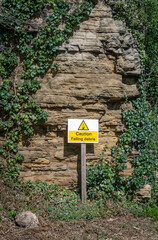 Beware of falling rock warning sign with triangle picture and alert message in yellow and black.  Stood in front of cliff face with rock on the ground.
